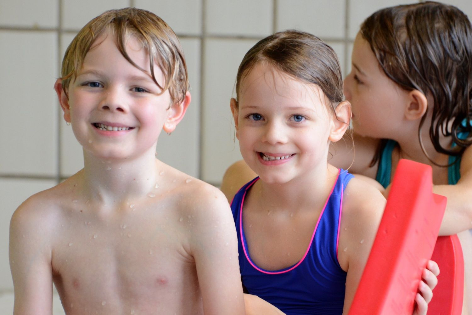 Drei lachende Kinder mit Schwimmbrett am Beckenrand